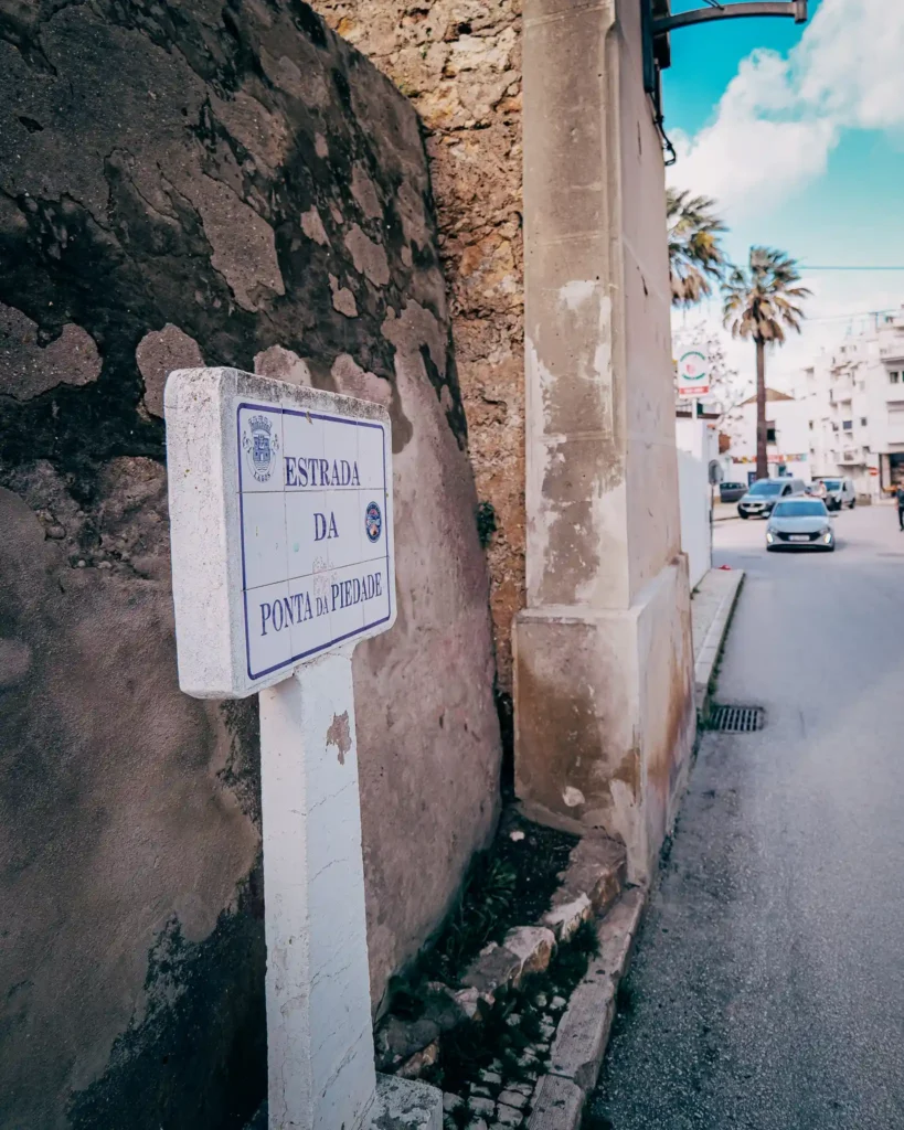Rue étroite à Lagos, Portugal, avec panneau "Estrada da Ponta da Piedade" et mur en pierre sous ciel bleu.