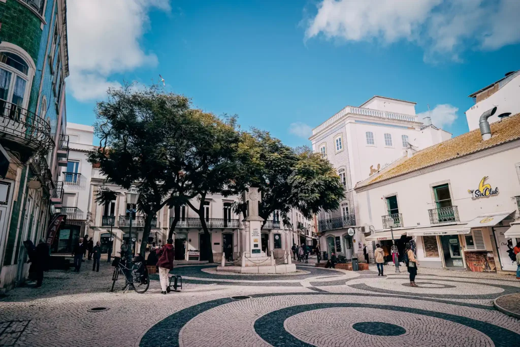 Place animée à Lagos, Portugal, avec des bâtiments historiques, arbres et passants sous un ciel bleu.