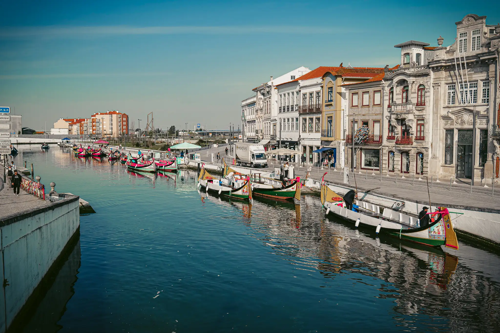 Aveiro canal with colorful moliceiros, historic buildings in the background, bright blue sky.