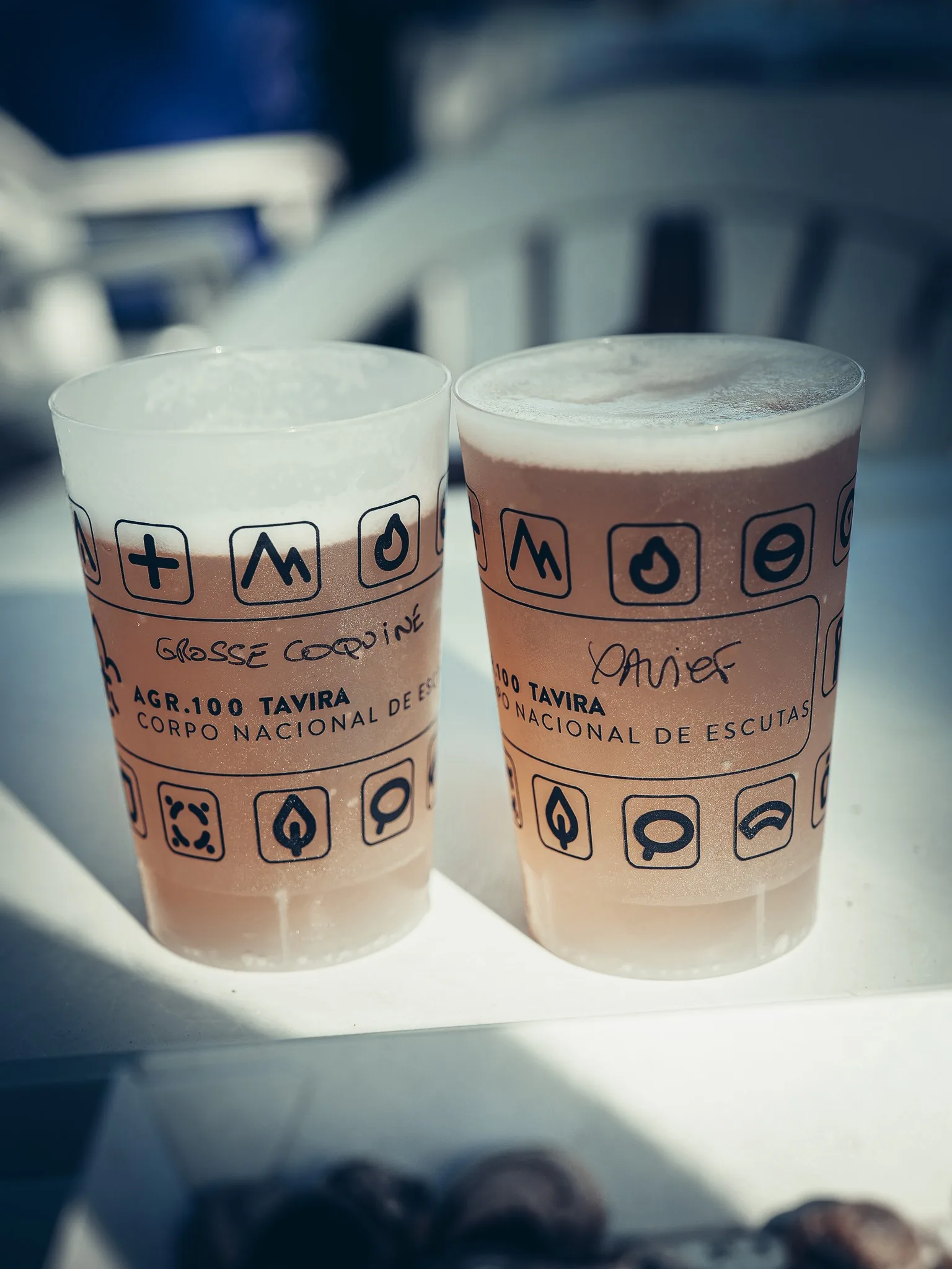 Two beer cups placed on a white plastic table, with inscriptions related to Tavira and the Corpo Nacional de Escutas, during an outdoor event in Tavira, Portugal. The style is marked for Caroline's beer glass "Grosse Coquine" and for Xavier's glass "Xavier".
