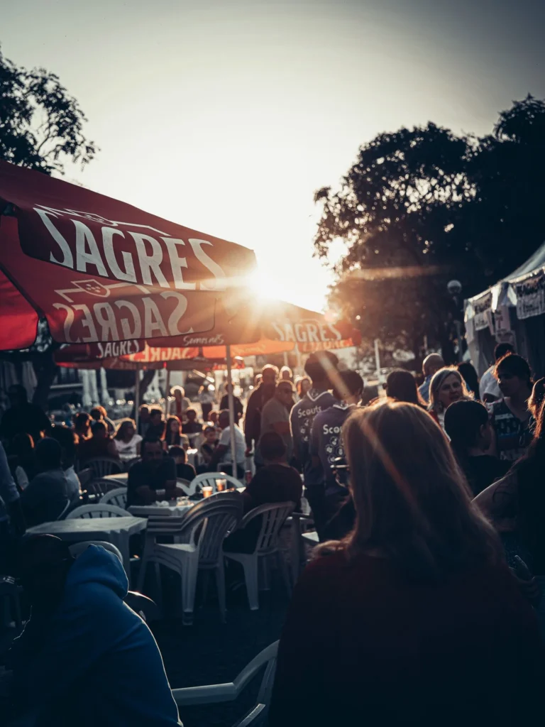 Lively terrace during an outdoor festival in Tavira at sunset, with many participants sitting under red Sagres umbrellas.