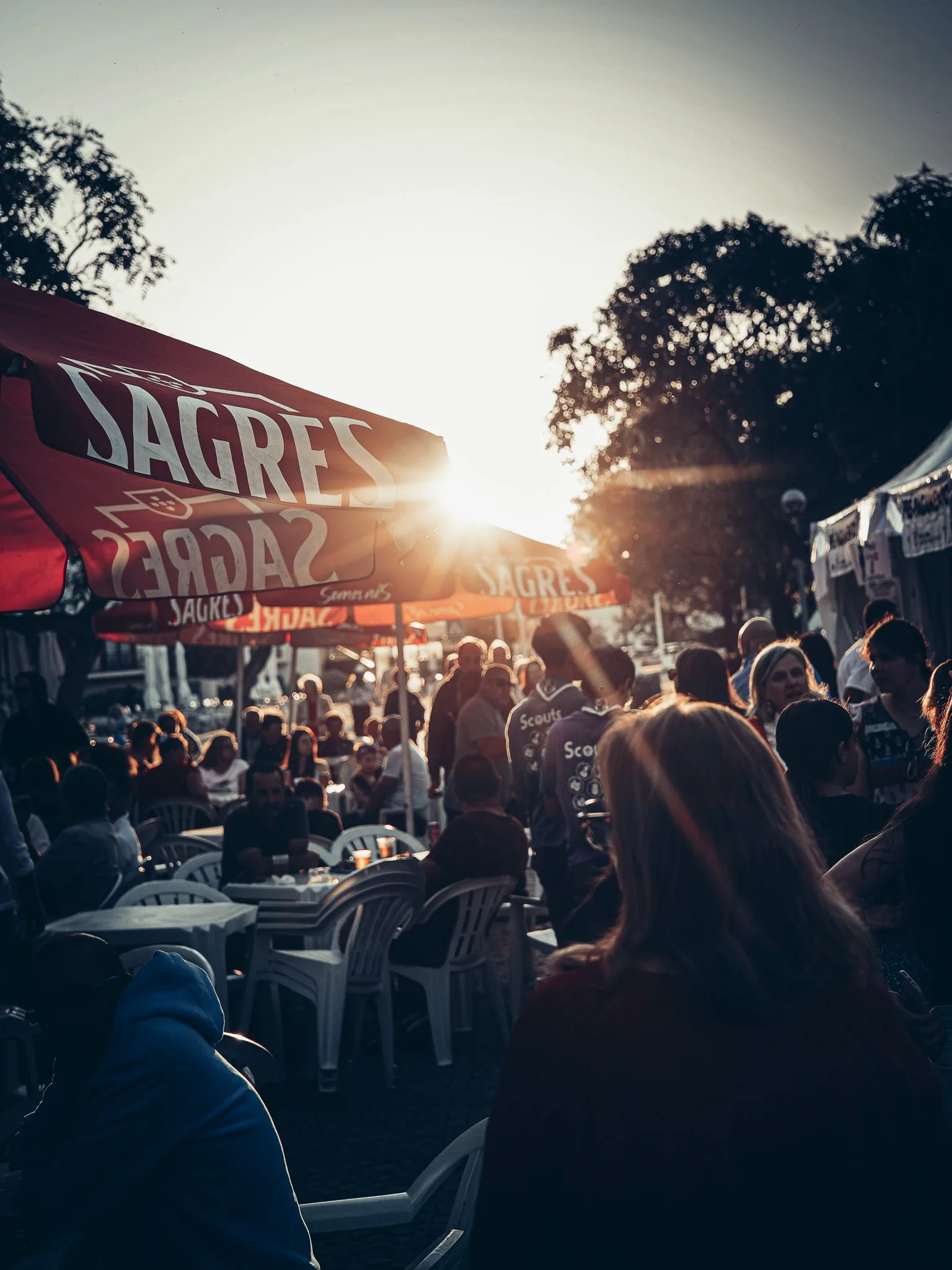 Lively terrace during an outdoor festival in Tavira at sunset, with many participants sitting under red Sagres umbrellas.