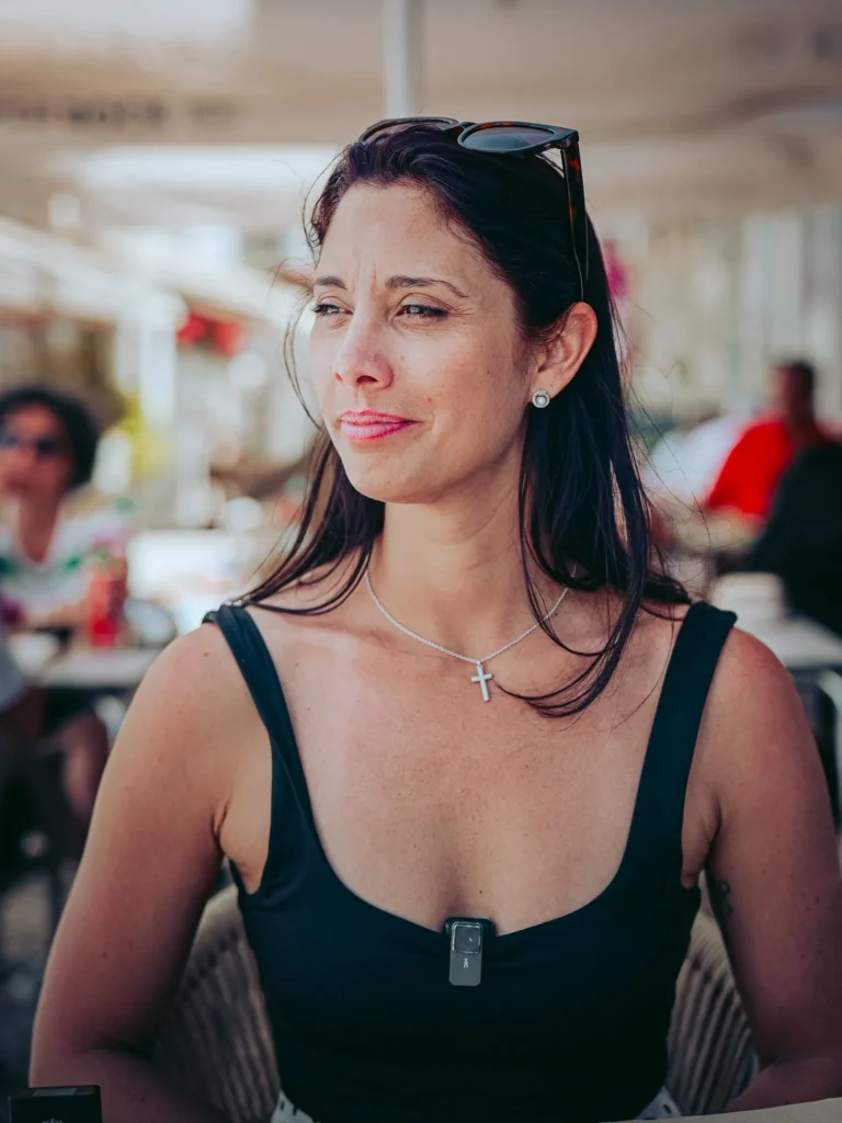 Caroline sitting on the terrace of a restaurant in Portimao, wearing a black tank top and a necklace with a cross, under natural light.