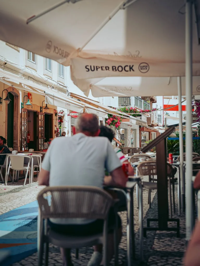 Xavier is sitting at an outdoor restaurant terrace under a large Super Bock umbrella, in a lively and flower-filled pedestrian street.