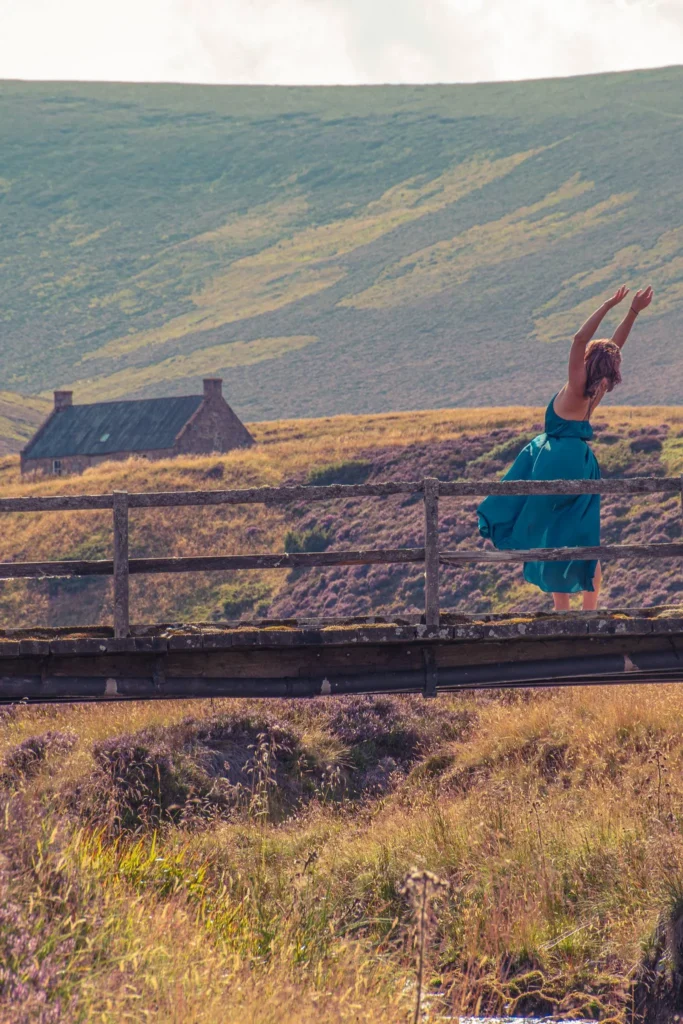Caroline in a blue dress dances on a wooden bridge in the heart of the moors of Cairngorms National Park