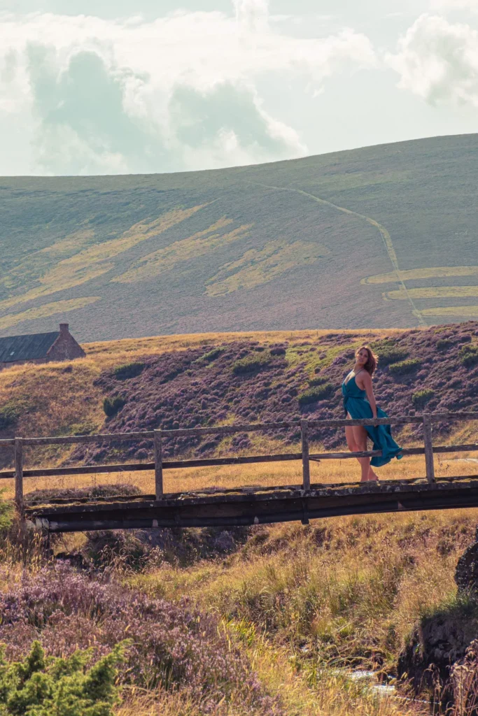 Caroline in a blue dress on a wooden bridge in the hilly landscape of the Cairngorms