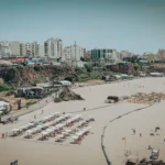 Large sandy beach in Portimao with aligned umbrellas, a few holidaymakers, and buildings in the background, under a clear sky in the Algarve at the end of May.