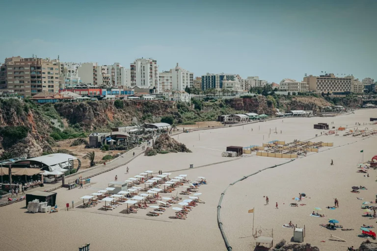 Large sandy beach in Portimao with aligned umbrellas, a few holidaymakers, and buildings in the background, under a clear sky in the Algarve at the end of May.