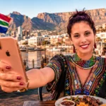 Caroline at V&A Waterfront, Cape Town, holding a smartphone with Table Mountain in the background.