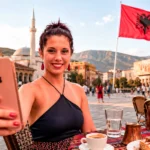 Caroline taking a selfie with her smartphone on a terrace in Tirana, Albania, with the Albanian flag in the background.
