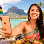 Caroline smiling with her smartphone in front of Le Morne Brabant, Mauritius, Mauritian flag waving.