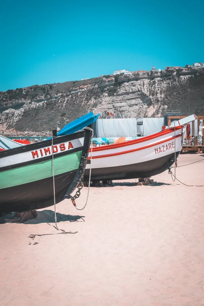 Traditional fishing boats on Nazaré beach, with the cliffs in the background under a blue sky in summer.