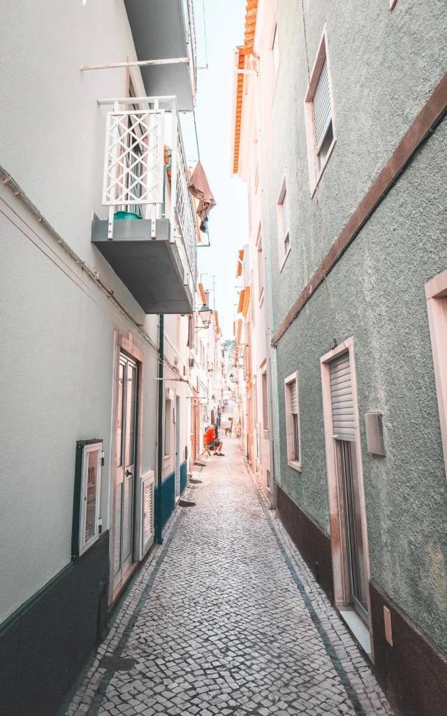 Narrow cobbled alley lined with colorful houses in Nazaré, Portugal, in summer 2023. No clearly visible passerby in the foreground.