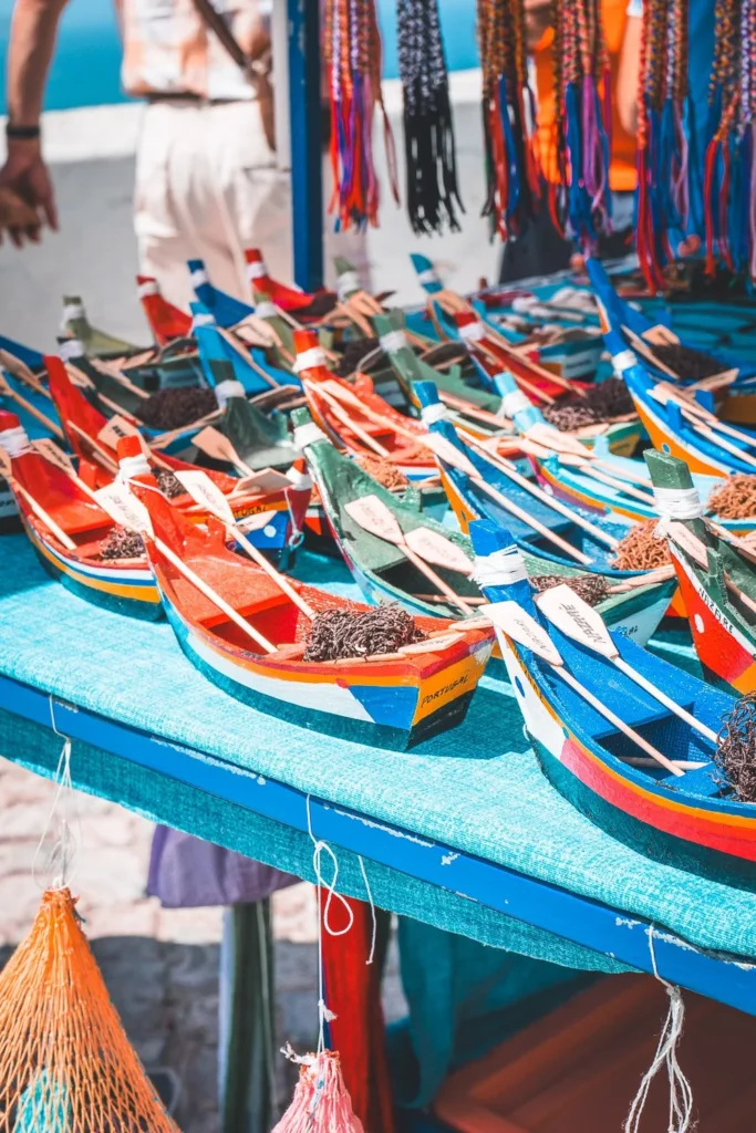 Souvenir stalls in Nazaré with colorful models of traditional Portuguese fishing boats displayed on an outdoor table in summer.