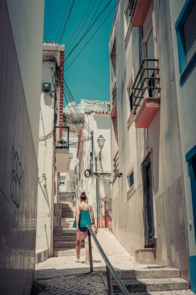 Caroline climbs a narrow staircase lined with white houses in a typical alley of Nazaré under a blue summer sky.