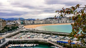 Aerial view of La Concha Bay in San Sebastián: the marina and its boats in the foreground, the shell-shaped beach, the Ferris wheel and the city in the background, autumn plane tree branch and sun rays piercing a cloudy sky, November 2021.