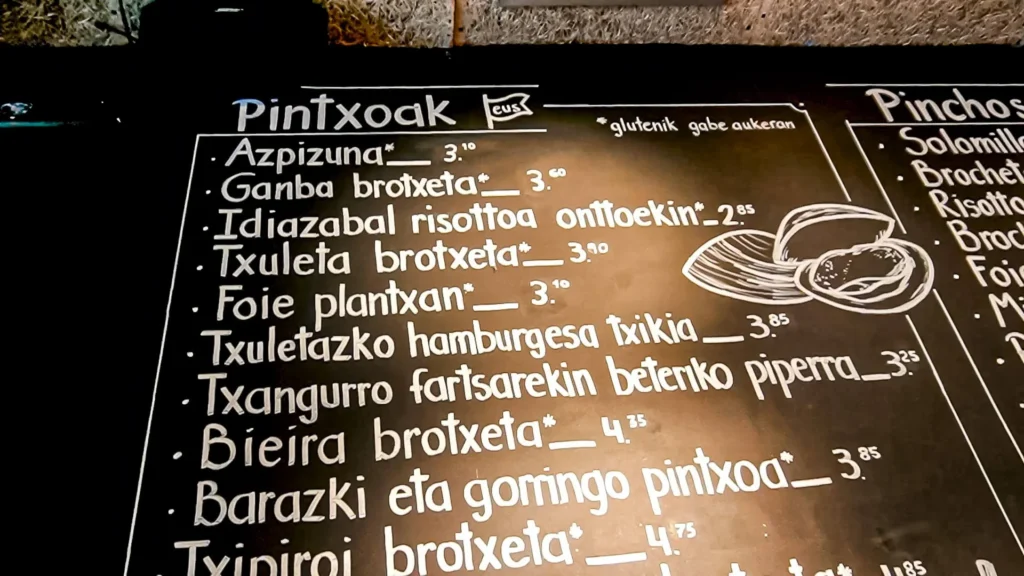 Handwritten slate from a pintxos bar in San Sebastián listing the "Pintxoak" in Basque (txuleta brotxeta, foie plantxan, txangurro, bieira) at around 3 € each, with gluten-free options, November 2021.