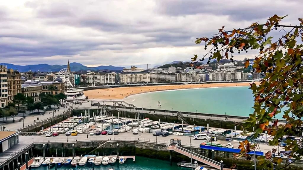 Aerial view of La Concha Bay in San Sebastián: the marina and its boats in the foreground, the shell-shaped beach, the Ferris wheel and the city in the background, autumn plane tree branch and sun rays piercing a cloudy sky, November 2021.