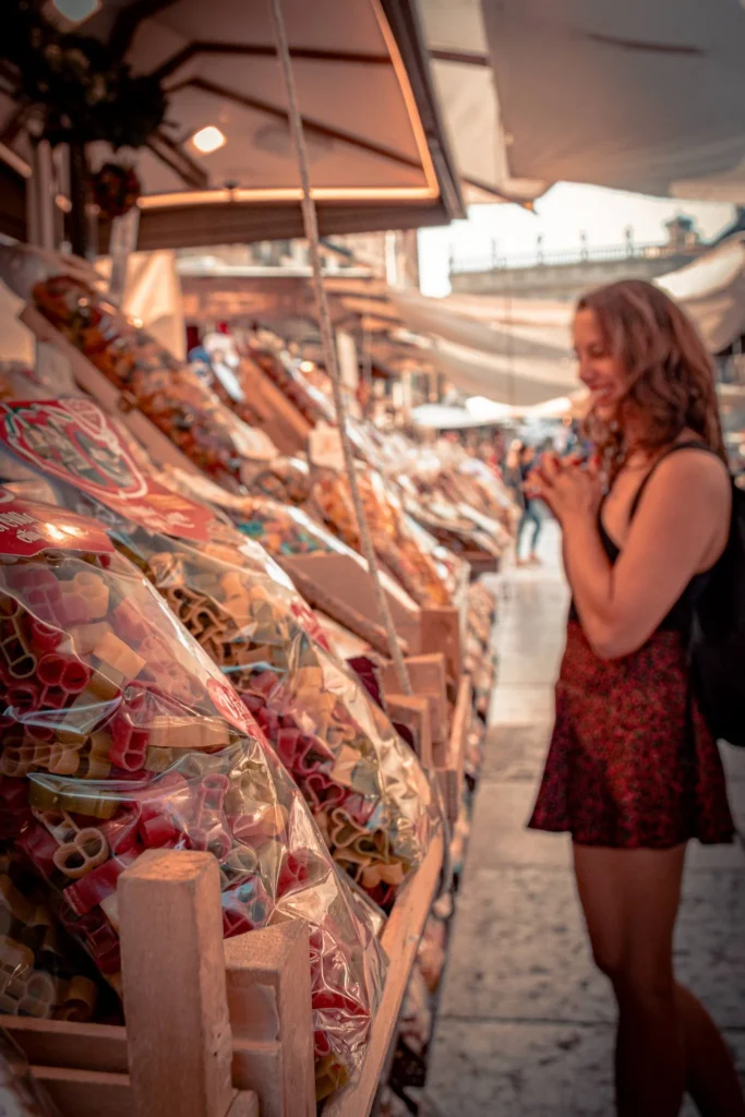 Caroline observes a market stall of colorful pasta bags at Piazza delle Erbe in Verona.