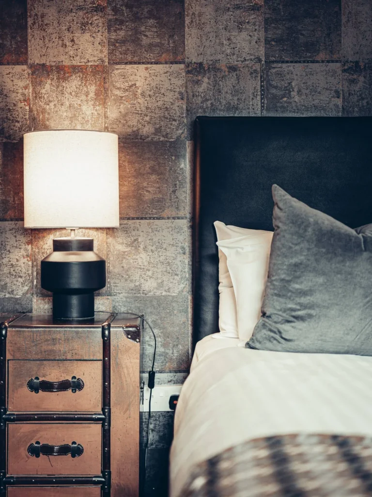 Detail of a room at the Allan Park Hotel in Stirling, featuring a wooden bedside table, a lit lamp, and a padded headboard, with a warm and contemporary atmosphere.