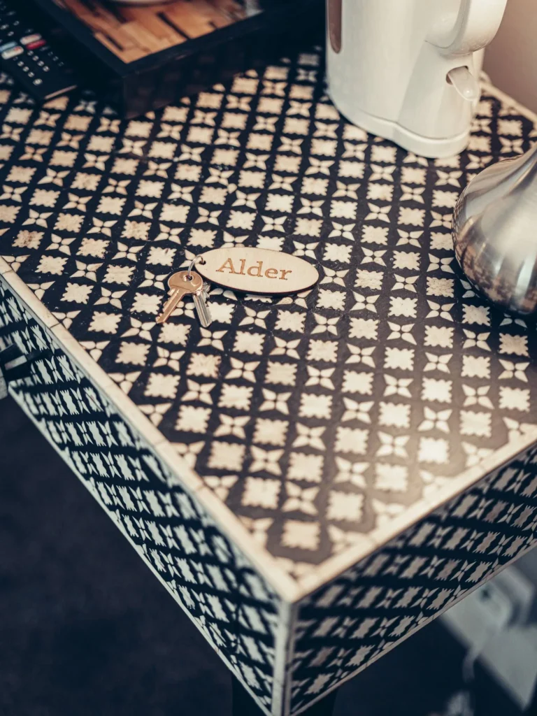 Detail of a bedside table decorated with a geometric pattern, on which rest room keys with an 'Alder' keychain, a white kettle, and a metal vase, in a room at the Allan Park Hotel in Stirling in August 2026.