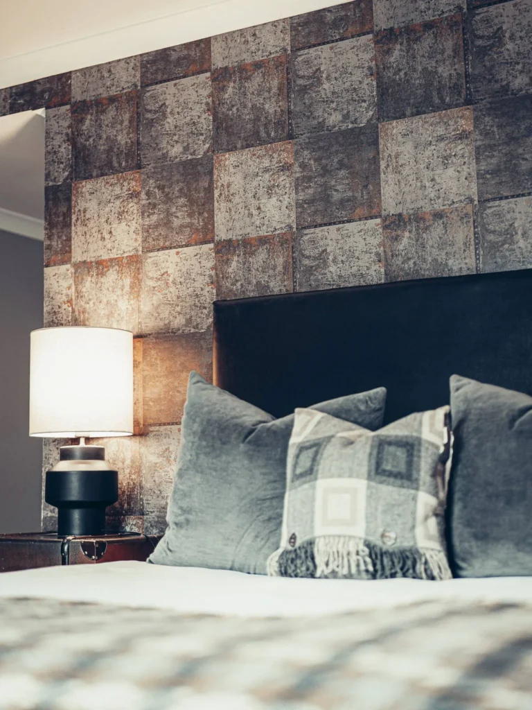 Detail of a room at the Allan Park Hotel in Stirling, featuring a dark headboard, grey cushions, and a lit bedside lamp in front of a geometric patterned wall. Cozy and contemporary atmosphere in Scotland in mid-August.
