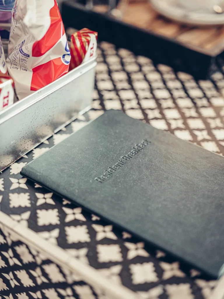 Notebook titled 'Resident Breakfast' placed on a patterned table, next to a tray containing packets of biscuits or snacks in a room at the Allan Park Hotel in Stirling in August 2026.