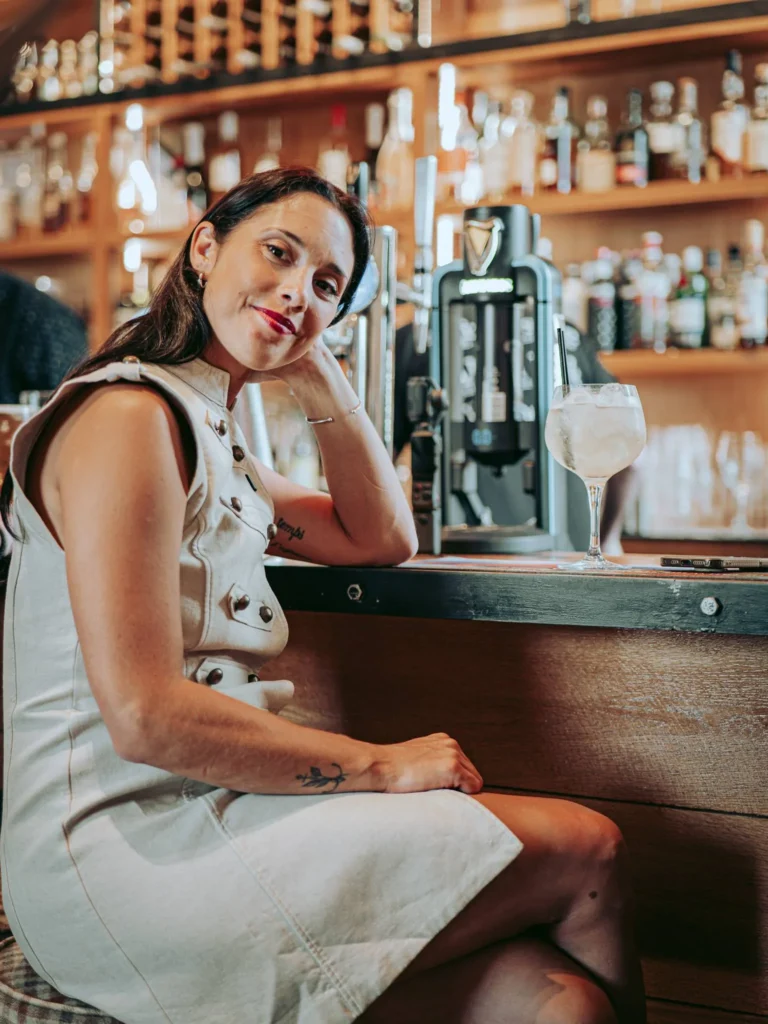 Caroline sitting at the bar of the Allan Park Hotel in Stirling, Scotland, in August 2026, a cocktail in front of her and bottles in the background.