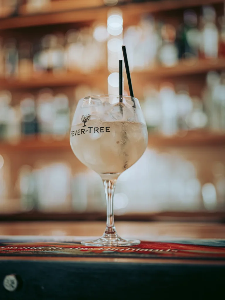 A large Fever-Tree cocktail glass filled with ice is placed on the bar counter of the Allan Park Hotel in Stirling, Scotland, with blurred bottles in the background.