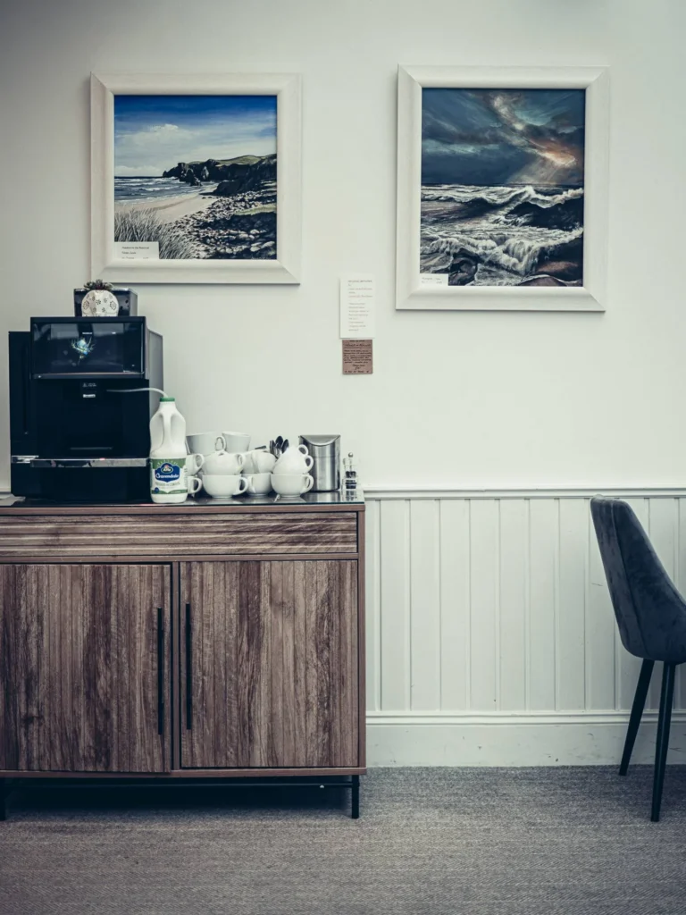 Coffee corner in a room at the Allan Park Hotel in Stirling, with coffee machine, cups, milk, and two paintings depicting coastal landscapes hanging on the wall.