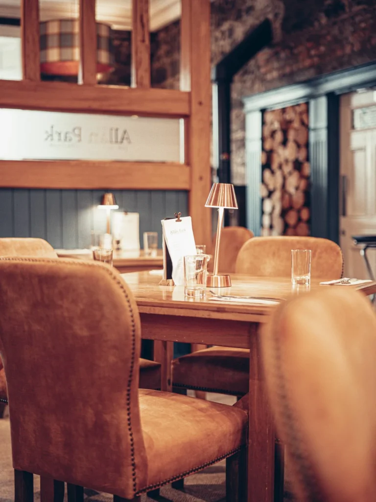 Restaurant with a warm atmosphere at the Allan Park Hotel in Stirling, featuring wooden furniture and beige velvet chairs, with no customers present.