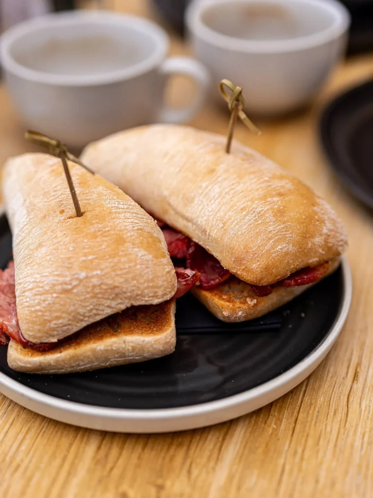 Two ciabatta bread sandwiches filled with cured meat are placed on a black plate, with coffee cups in the background on a wooden table in a room at the Allan Park Hotel in Stirling in August 2026.
