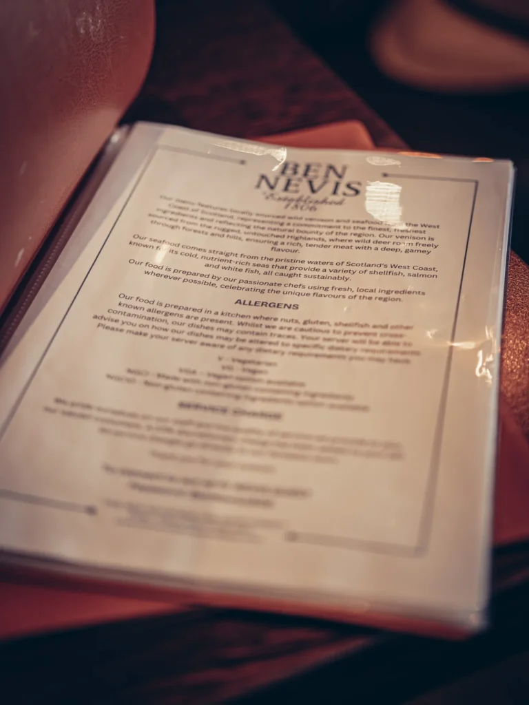 Menu of the Ben Nevis Bar placed on a wooden table in a warm atmosphere in Fort William, in the Scottish Highlands, on August 18, 2026.