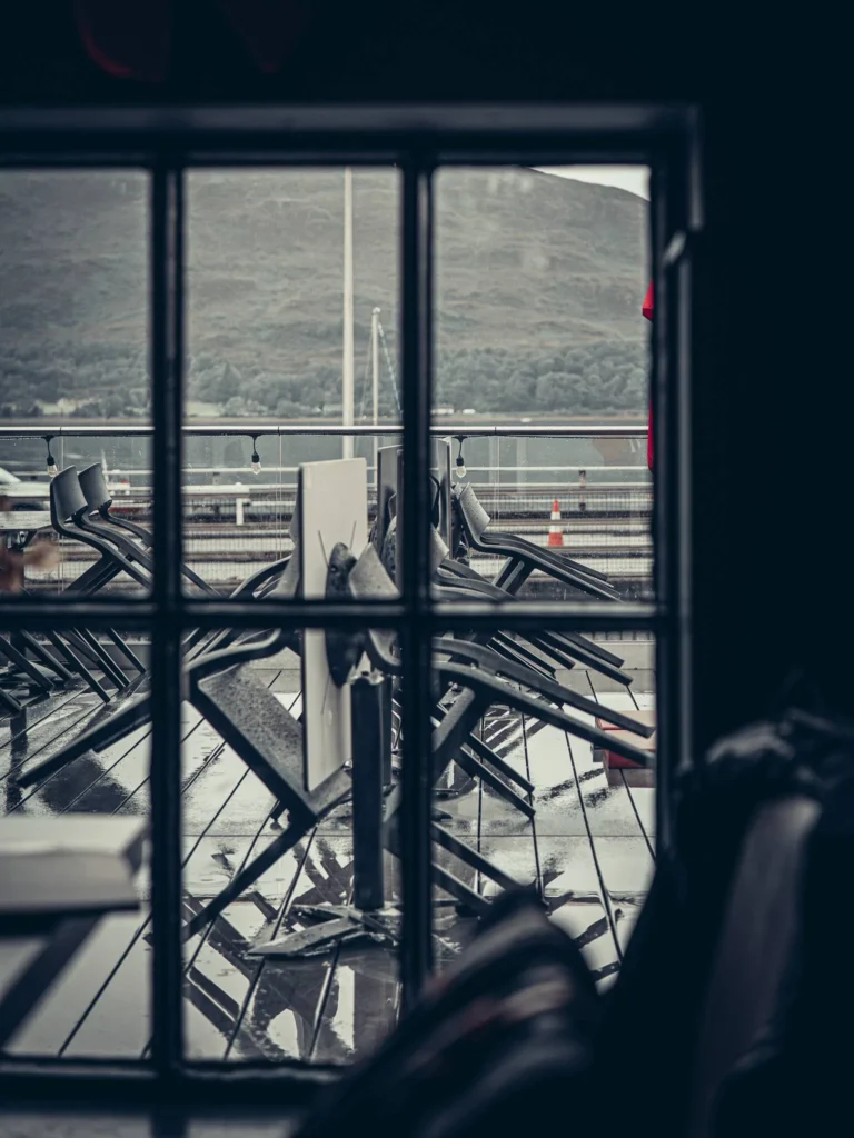 View from inside the Ben Nevis Bar onto a wet outdoor terrace, with stacked chairs and the Highlands mountains in the background.