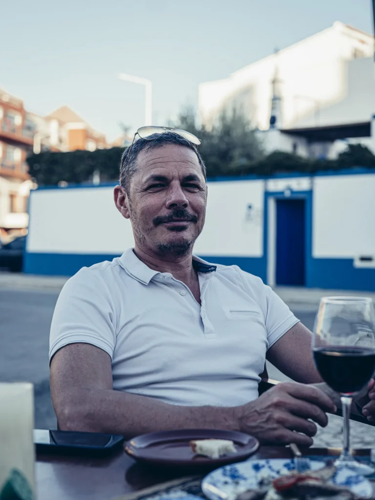 Xavier seated on the terrace at Bodeguita Salinas in Tavira, a glass of red wine and tapas under the evening light, with white and blue facades in the background.