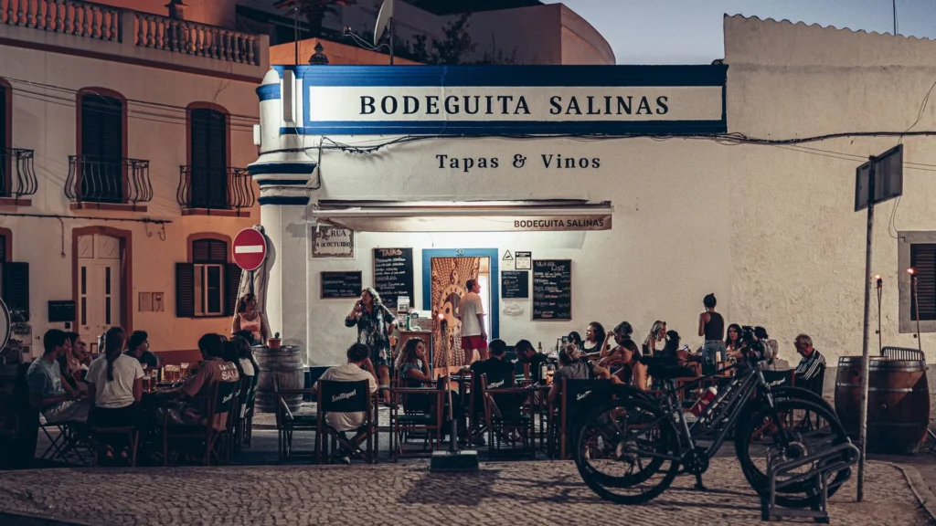 Lively terrace of Bodeguita Salinas in Tavira on a summer evening: the tapas and wine bar occupies the car-free street, with several groups seated outside.