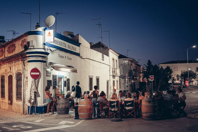 Lively terrace of the Bodeguita Salinas in Tavira on a summer evening: groups seated around wine barrels turned into high tables, sign "Tapas & Vinos," light from the street lamps.