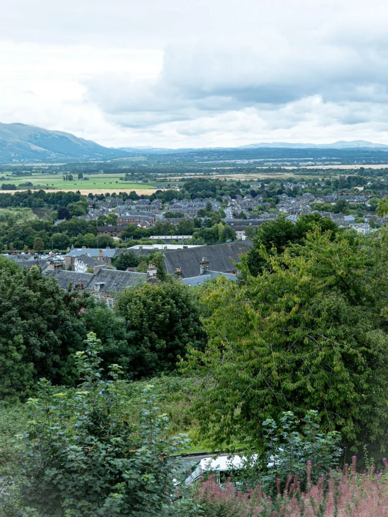 A panorama over the town of Stirling from the heights of the castle, stone rooftops among trees with hills on the horizon under a cloudy sky.