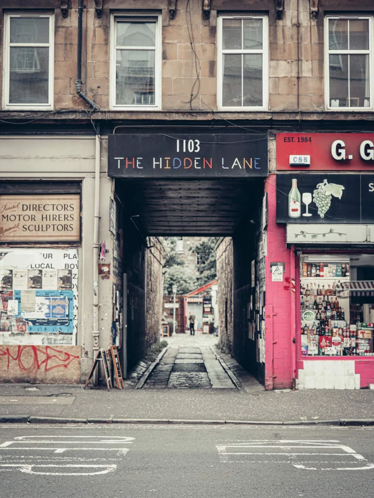 The entrance porch of The Hidden Lane, at 1103 Argyle Street in Finnieston (Glasgow): the sign “1103 THE HIDDEN LANE” sits above a long passage under the building, wide enough for a car, which opens onto the courtyard of studios. On the right, the wine merchant GG Brothers Liquor; on the left, an old painted shopfront reading “Funeral Directors / Motor Hirers / Monumental Sculptors”.