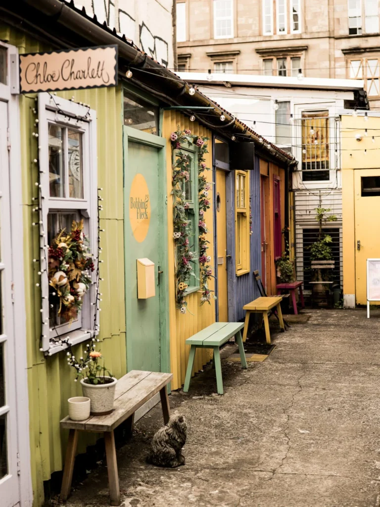 Colourful studio facades on The Hidden Lane in Glasgow, hung with fairy lights and flowers, with wooden benches and potted plants. The decoration cuts straight through the mineral monotony of Glasgow, a city Xavier finds “beautiful in its sadness”.
