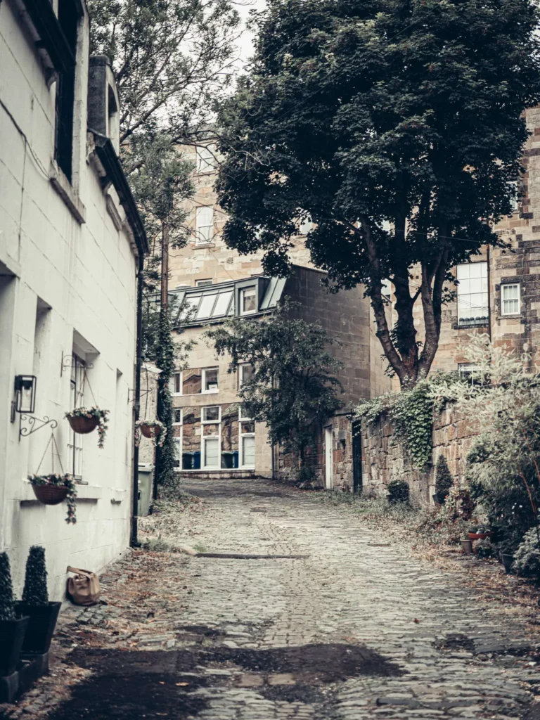 Cobbled lane lined with blond sandstone houses and greenery, under the changing Glasgow sky.