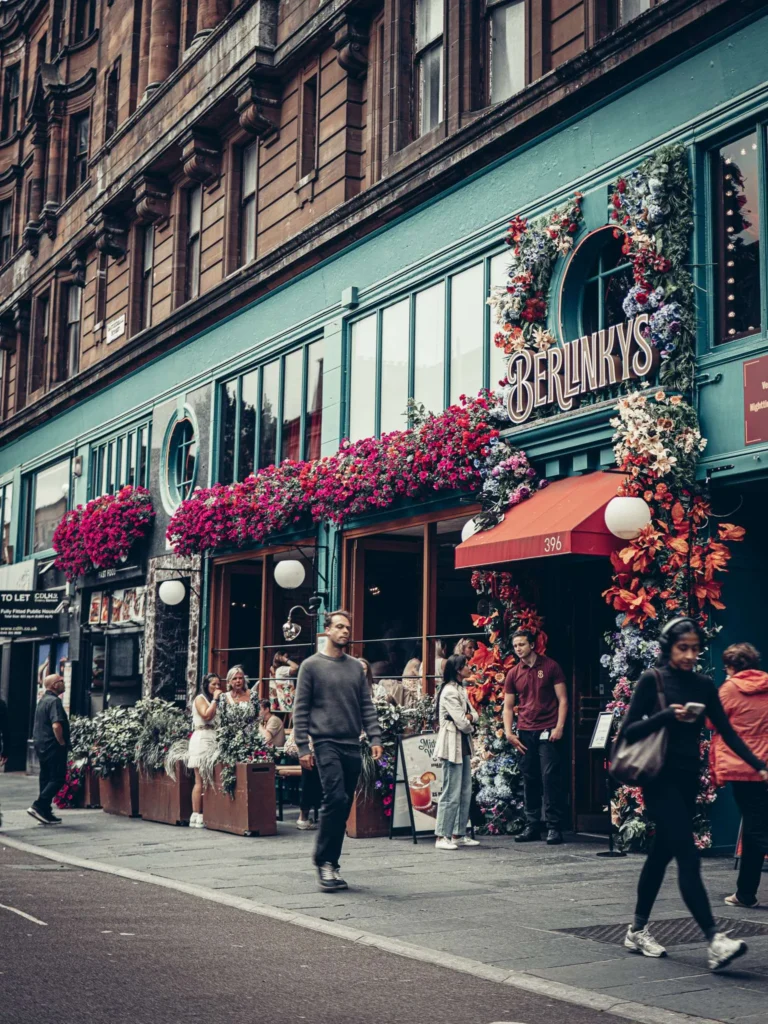 Facade of a café overflowing with flowers on a busy street in Glasgow, in front of a Victorian blond sandstone building.