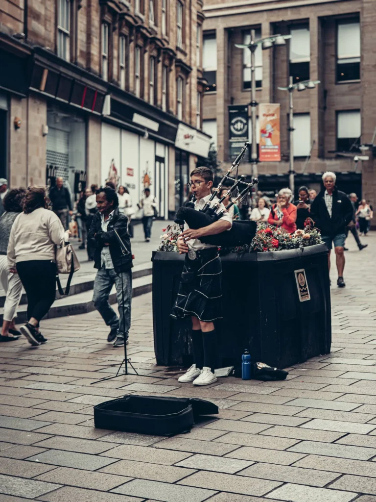 Bagpiper in a kilt on a cobbled shopping street in the centre of Glasgow, between the blond sandstone buildings.