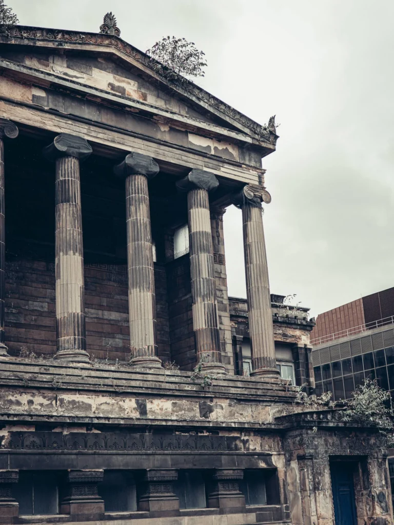 Neoclassical sandstone facade with columns in Glasgow, marked by wear, with vegetation growing on the cornice.