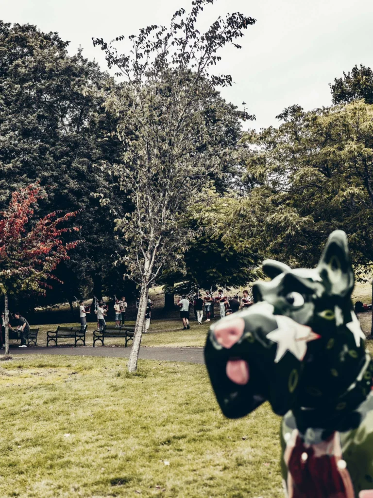 A decorated dog sculpture in the foreground of a park in Glasgow, with the pipers playing behind.