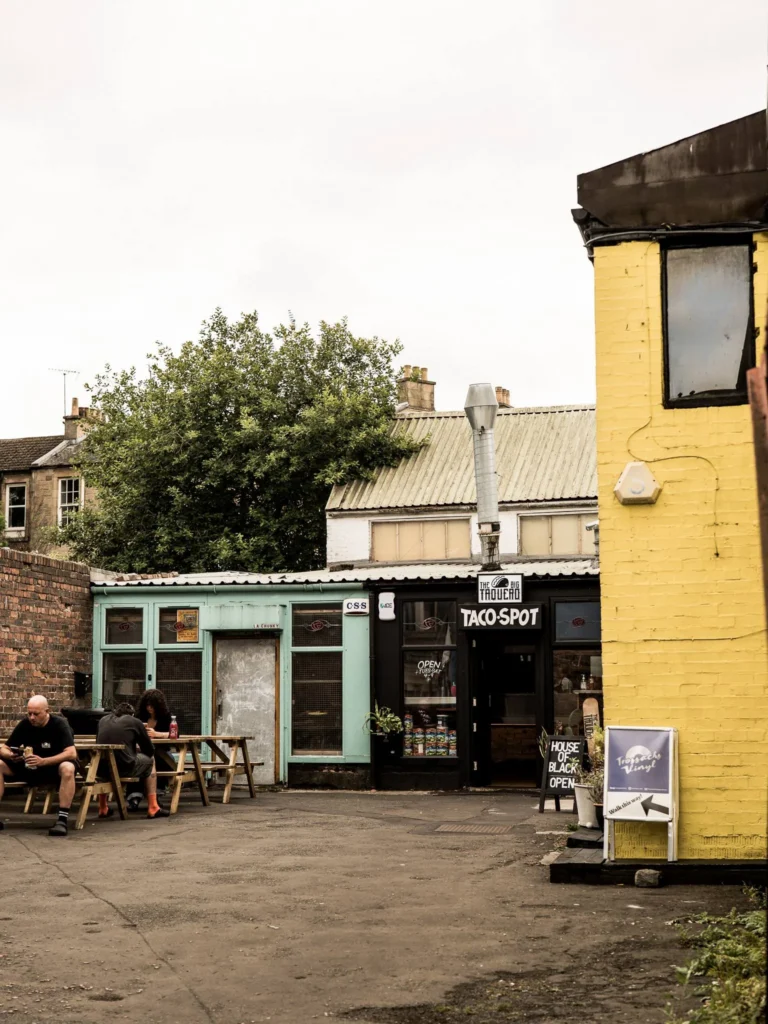 The Hidden Lane courtyard in Glasgow: colorful facades, wooden benches, and a few people sitting outside in overcast weather. We pass under a building and emerge into what Xavier describes as a neighborhood within a neighborhood.
