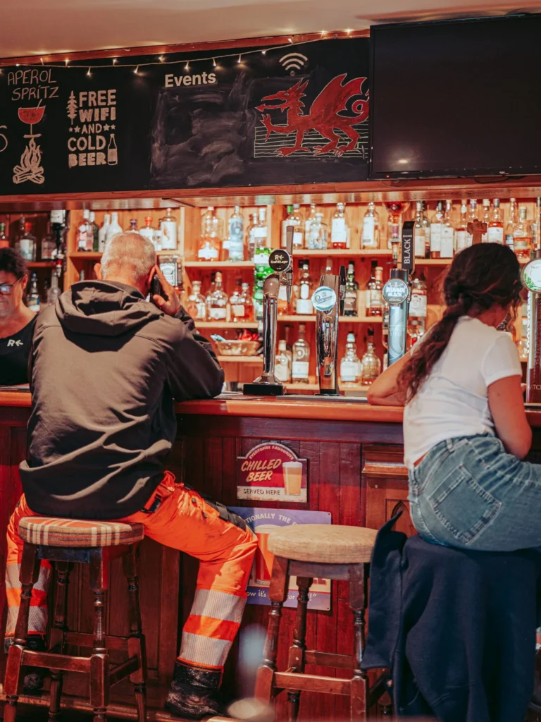 Two people at the counter of a warm bar, surrounded by bottles and beer pumps, in a Scottish pub setting.