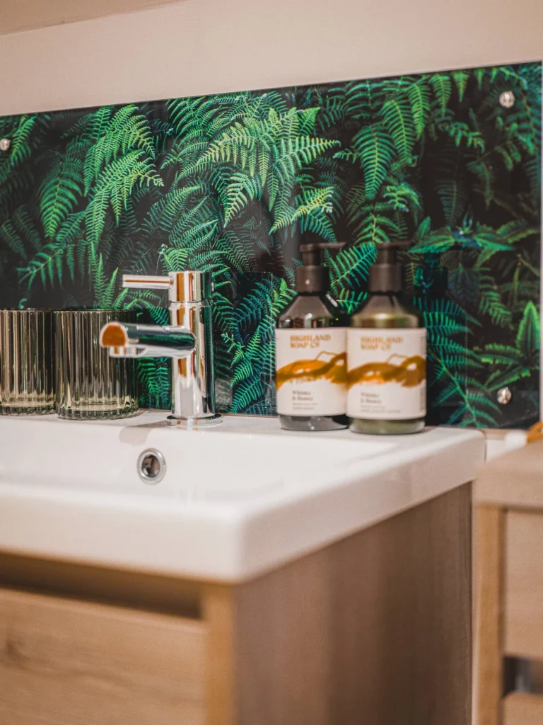 Modern bathroom basin with chrome tap, two glasses, and Highland Soap Co toiletries in front of a decorative panel featuring fern patterns. Natural and tidy atmosphere in accommodation in the Scottish Highlands.