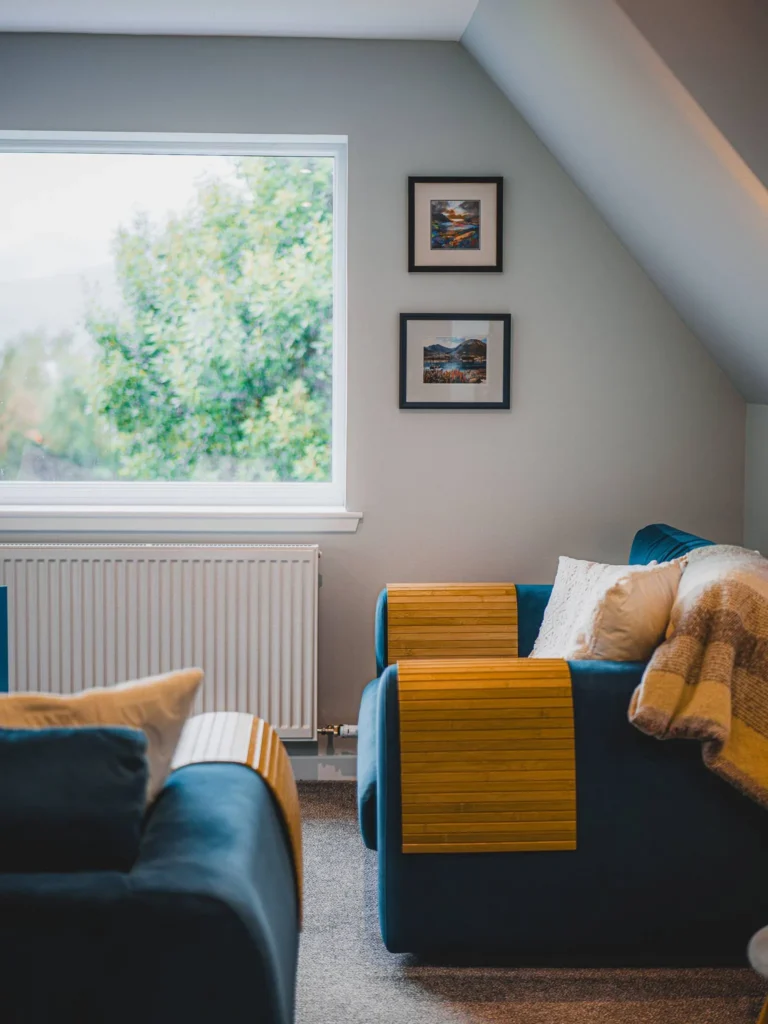 Cosy sitting room of a house in Glencoe in the Highlands, with two blue sofas, a large window overlooking the greenery, and paintings hanging on the wall.
