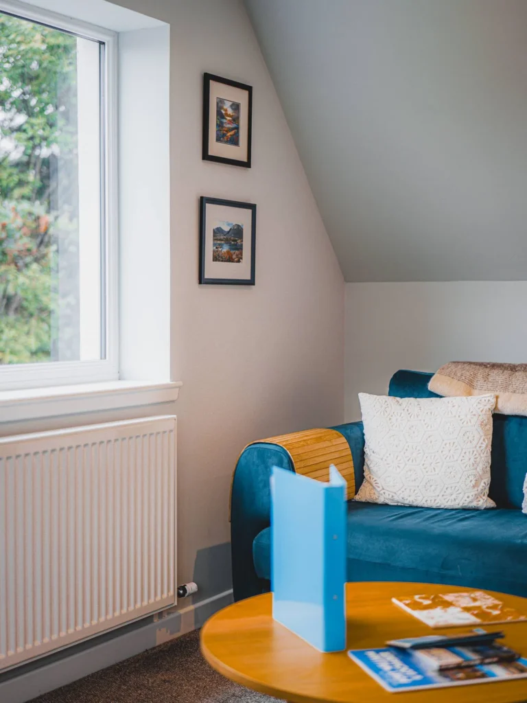 Bright living area of an accommodation in Glencoe in the Scottish Highlands, featuring a blue sofa, knitted cushion, coffee table, and a view of the outdoor greenery.